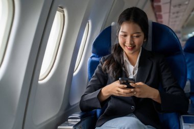 İş kadını mesajlaşmak için akıllı telefonu kullanıyor ve tatil zamanı yaşam tarzı sırasında uçuştaki iş seyahatlerinde pencerelerin yanında otururken finans sektörü raporlarını okumak için çalışıyor..