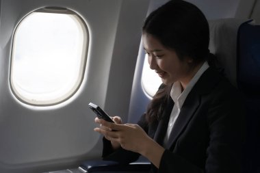 İş kadını mesajlaşmak için akıllı telefonu kullanıyor ve tatil zamanı yaşam tarzı sırasında uçuştaki iş seyahatlerinde pencerelerin yanında otururken finans sektörü raporlarını okumak için çalışıyor..