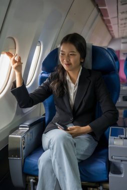 İş kadını kulübelerin pencerelerini açıyor gökyüzü manzarasına mutlulukla bakıyor ve akıllı telefon üzerinde çalışıyor. Tatil sırasında uçakta iş seyahati sınıfının pencerelerinin yanında oturuyor..