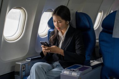 İş kadınları müzik dinlemek için bluetooth kulaklık takarlar ve uçaktaki uçuş işi seyahatleri sırasında pencerelerin yanında otururken akıllı telefondan yeni bir girişimin finansmanı hakkındaki iş raporlarını kontrol ederler..