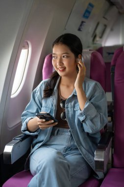 Asyalı gezgin kadınlar müzik dinlemek için bluetooth kulaklık takıyor ve uçuş sırasında camların yanında tatil yaşam tarzıyla seyahat ederken akıllı telefondan sohbet mesajları yazıyorlar..