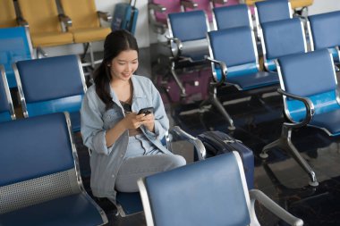 Asyalı gezgin kadın sosyal medyada sörf yapıyor ve havaalanındaki bankta oturmuş business travel class uçuşuyla seyahat etmeyi beklerken akıllı telefondan sohbet mesajları yazıyor..