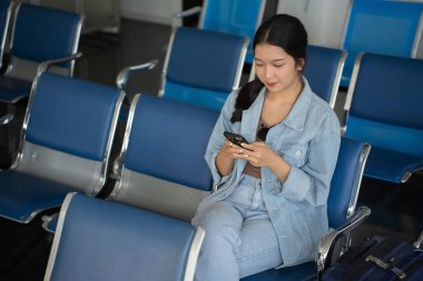 Asyalı gezgin kadın sosyal medyada sörf yapıyor ve havaalanındaki bankta oturmuş business travel class uçuşuyla seyahat etmeyi beklerken akıllı telefondan sohbet mesajları yazıyor..