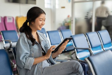 Asyalı gezgin kadın sosyal medyada sörf yapmak ve sohbet mesajları yazmak için tablet kullanırken terminal havaalanında bankta oturup iş seyahati sınıfı uçuşuyla tatil yaşam tarzını beklemektedir..