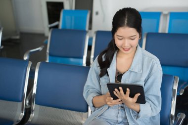 Asyalı gezgin kadın sosyal medyada sörf yapmak ve sohbet mesajları yazmak için tablet kullanırken terminal havaalanında bankta oturup iş seyahati sınıfı uçuşuyla tatil yaşam tarzını beklemektedir..