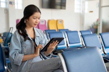 Asyalı gezgin kadın sosyal medyada sörf yapmak ve eğlenmek için tablet kullanıyor. Terminal havaalanında bankta oturmuş business travel class uçuşuyla seyahat etmeyi beklerken..