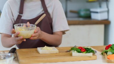 Asyalı kadınlar ellerinde bir kase haşlanmış yumurta salatası tutarken sandviç yapmak için ekmek ve sebze koyarken kahvaltı hazırlamak ve evde sağlıklı yemek yapmak için gerekli malzemeleri hazırlıyorlar..