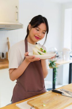 Asyalı kadınlar kaynamış yumurta salatası ve taze sebzeyle sandviç yaptıktan sonra ellerinde lezzetli bir tabak sandviç tutarak kahvaltı hazırlıyorlar ve evde sağlıklı bir yaşam tarzı yaratıyorlar..