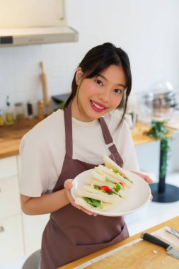 Asyalı kadınlar kaynamış yumurta salatası ve taze sebzeyle sandviç yaptıktan sonra ellerinde lezzetli bir tabak sandviç tutarak kahvaltı hazırlıyorlar ve evde sağlıklı bir yaşam tarzı yaratıyorlar..