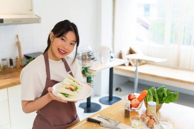 Asyalı kadınlar kaynamış yumurta salatası ve taze sebzeyle sandviç yaptıktan sonra ellerinde lezzetli bir tabak sandviç tutarak kahvaltı hazırlıyorlar ve evde sağlıklı bir yaşam tarzı yaratıyorlar..