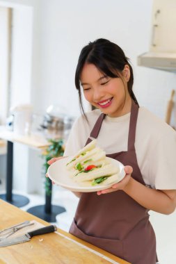 Asyalı kadınlar kaynamış yumurta salatası ve taze sebzeyle sandviç yaptıktan sonra ellerinde lezzetli bir tabak sandviç tutarak kahvaltı hazırlıyorlar ve evde sağlıklı bir yaşam tarzı yaratıyorlar..