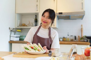 Asyalı kadınlar kaynamış yumurta salatası ve taze sebzeyle sandviç yaptıktan sonra ellerinde lezzetli bir tabak sandviç tutarak kahvaltı hazırlıyorlar ve evde sağlıklı bir yaşam tarzı yaratıyorlar..