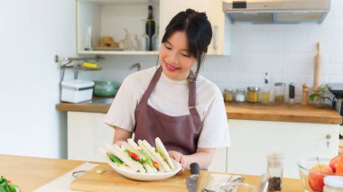 Asyalı kadınlar kaynamış yumurta salatası ve taze sebzeyle sandviç yaptıktan sonra ellerinde lezzetli bir tabak sandviç tutarak kahvaltı hazırlıyorlar ve evde sağlıklı bir yaşam tarzı yaratıyorlar..