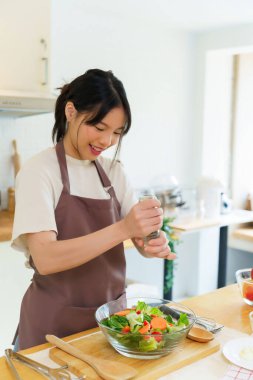Asyalı kadınlar kahvaltı yemeği ve sebze salatası hazırlamak için malzemeyi karıştırırken aynı zamanda evde, mutfakta ev yapımı sağlıklı yiyecekler yapmak için cam bir kase sebzeye biber ekliyorlar..
