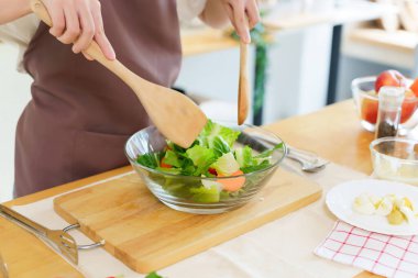 Asyalı kadınların elleri, cam kasede malzeme ve taze sebze dilimlerini karıştırmak için tahta kaşık kullanırken kahvaltılık yemek ve sebze salatası pişirirken sağlıklı bir yaşam tarzı yaratıyor..