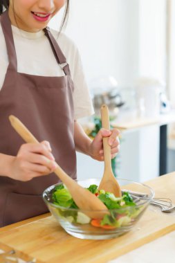 Asyalı kadınlar cam kasede taze sebze ve malzemeyi karıştırmak için tahta kaşık kullanırken kahvaltılık yemek ve sebze salatası pişirirken aynı zamanda ev yapımı sağlıklı yiyecek yaşam tarzı yaratıyorlar..