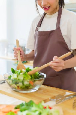 Asyalı kadınlar cam kasede taze sebze ve malzemeyi karıştırmak için tahta kaşık kullanırken kahvaltılık yemek ve sebze salatası pişirirken aynı zamanda ev yapımı sağlıklı yiyecek yaşam tarzı yaratıyorlar..
