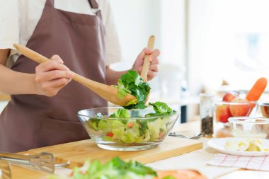 Asyalı kadınlar cam kasede taze sebze ve malzemeyi karıştırmak için tahta kaşık kullanırken kahvaltılık yemek ve sebze salatası pişirirken aynı zamanda ev yapımı sağlıklı yiyecek yaşam tarzı yaratıyorlar..