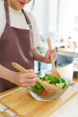 Asyalı kadınlar cam kasede taze sebze ve malzemeyi karıştırmak için tahta kaşık kullanırken kahvaltılık yemek ve sebze salatası pişirirken aynı zamanda ev yapımı sağlıklı yiyecek yaşam tarzı yaratıyorlar..