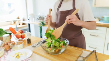 Asyalı kadınlar cam kasede taze sebze ve malzemeyi karıştırmak için tahta kaşık kullanırken kahvaltılık yemek ve sebze salatası pişirirken aynı zamanda ev yapımı sağlıklı yiyecek yaşam tarzı yaratıyorlar..