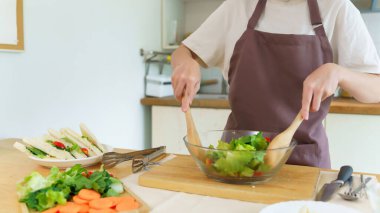 Asyalı kadınlar cam kasede taze sebze ve malzemeyi karıştırmak için tahta kaşık kullanırken kahvaltılık yemek ve sebze salatası pişirirken aynı zamanda ev yapımı sağlıklı yiyecek yaşam tarzı yaratıyorlar..