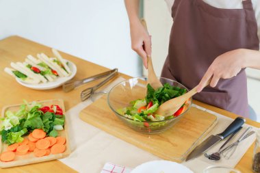 Asyalı kadınlar cam kasede taze sebze ve malzemeyi karıştırmak için tahta kaşık kullanırken kahvaltılık yemek ve sebze salatası pişirirken aynı zamanda ev yapımı sağlıklı yiyecek yaşam tarzı yaratıyorlar..