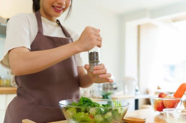 Asyalı kadınlar kahvaltı yemeği ve sebze salatası hazırlamak için malzemeyi karıştırırken aynı zamanda evde, mutfakta ev yapımı sağlıklı yiyecekler yapmak için cam bir kase sebzeye biber ekliyorlar..