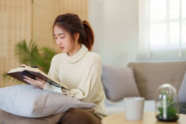 Rahat bir kanepede oturan genç Asyalı kadınlar rahatlarken düşünmek ve öğrenmek için kitap okuyorlar ve evde yavaş yaşam tarzıyla zaman geçirmek için aktivite yapıyorlar..