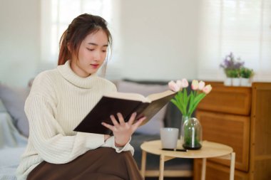 Kazak giyen genç Asyalı kadınlar kitap okuyup edebiyat hikayelerini öğrenirken koltukta rahatlamak ve evde yavaş bir yaşam tarzıyla vakit geçirmek için aktivite yaparken eğleniyorlar..