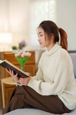 Süveter giyen genç Asyalı kadınlar kitap okurken ve edebiyat hikayelerini öğrenirken kanepede oturup dinlenirken ve evde yavaş yaşam tarzıyla zaman geçirmek için aktivite yaparken..