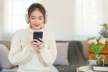 Kazaklı genç Asyalı kadınlar kulaklık takıyor ve akıllı telefondan müzik dinleyip eğlenirken evde yavaş bir yaşam tarzıyla vakit geçirmek için etkinlik yapıyorlar..