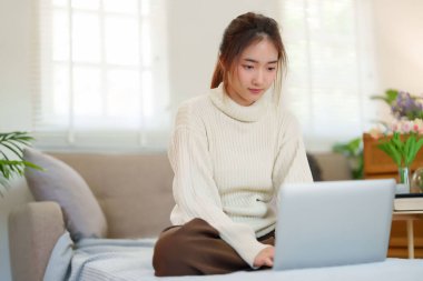 Kanepede oturan Asyalı genç kadınlar rahatlarken ve evde yavaş bir yaşam tarzıyla vakit geçirmek için aktivite yaparken dizüstü bilgisayarlarında sosyal medya eğlencesiyle eğleniyorlar..