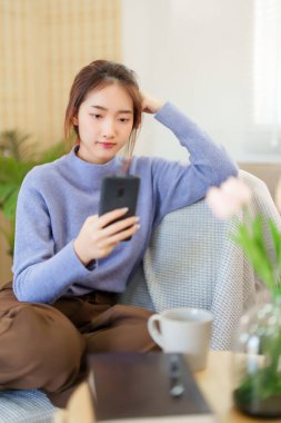 Genç Asyalı süveter giyen kadınlar sosyal medya ve eğlence sektöründe sörf yapmak için akıllı telefon kullanırken, aynı zamanda kanepede rahatlamak ve evde yavaş yaşam tarzıyla zaman geçirmek için aktivite yapmak için oturuyorlar..