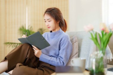 Genç Asyalı kadınlar kanepede oturup not defterine notlar ve seyahat günlüğü yazmak için yaslanırken, dinlenirken ve evde yavaş bir yaşam tarzıyla zaman geçirmek için etkinlik yaparken..