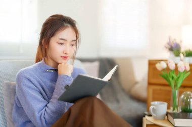 Kazaklı genç Asyalı kadınlar, evde yavaş bir yaşam tarzıyla vakit geçirmek ve rahatlamak için kanepede oturup etkinlik yaparken düşünceli ve bilgi eğitimi hakkında öğrenmek için bir kitap okuyorlar..