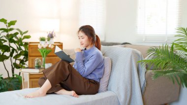 Kazaklı genç Asyalı kadınlar, evde yavaş bir yaşam tarzıyla vakit geçirmek ve rahatlamak için kanepede oturup etkinlik yaparken düşünceli ve bilgi eğitimi hakkında öğrenmek için bir kitap okuyorlar..