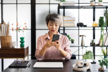 Kısa gri saçlı orta yaşlı bir iş kadını modern bir masada oturuyor, akıllı telefonuna odaklanmış, sakin ve profesyonel bir tavırla.