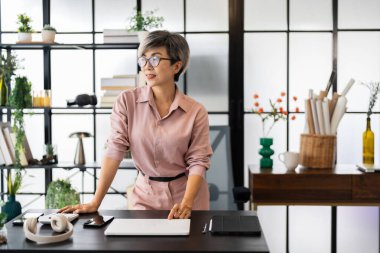 Orta yaşlı bir iş kadını masasında güven içinde duruyor, ellerini masaya dayayıp, modern bir ofis ortamında pencereden dışarı dikkatlice bakıyor.