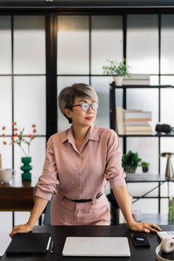 Kısa gri saçlı yaşlı iş kadını masasında güvenle duruyor, elleri masada dinleniyor, modern bir ofiste kararlılıkla ileri bakıyor..