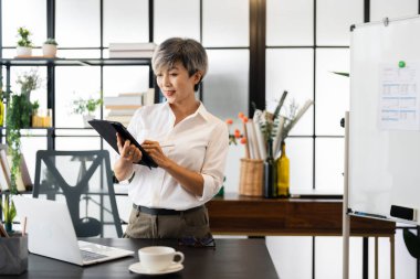 Kısa gri saçlı orta yaşlı bir iş kadını modern bir ofiste şık dekorasyon ve yeşillikle çevrili bir tabletle ayakta duruyor ve çalışıyor..