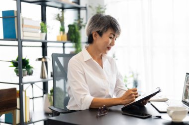 Orta yaşlı bir iş kadını, modern bir ofiste masasında oturmuş, odaklanmış ve konsantre olmuş bir tablet üzerinde çalışıyor..