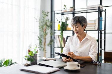 Orta yaşlı bir iş kadını, modern bir ofiste masasında oturmuş, odaklanmış ve konsantre olmuş bir tablet üzerinde çalışıyor..