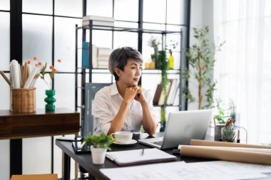 Bir bayan mimar masasında oturuyor, elinde bir akıllı telefon ve dizüstü bilgisayarına bakarken bir tasarım projesi tasarlıyor, yaratıcılığı ve odaklanmayı yansıtıyor..