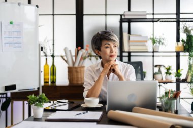 Bir bayan mimar masasında oturuyor, elinde bir akıllı telefon ve dizüstü bilgisayarına bakarken bir tasarım projesi tasarlıyor, yaratıcılığı ve odaklanmayı yansıtıyor..