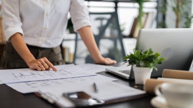 Bir kadın mimar masanın başında duruyor, iyi organize edilmiş ve modern ofis alanının planlarını gözden geçiriyor, detaylara odaklanma ve dikkat gösteriyor..