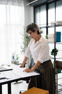 Bir kadın mimar masanın başında duruyor, iyi organize edilmiş ve modern ofis alanının planlarını gözden geçiriyor, detaylara odaklanma ve dikkat gösteriyor..