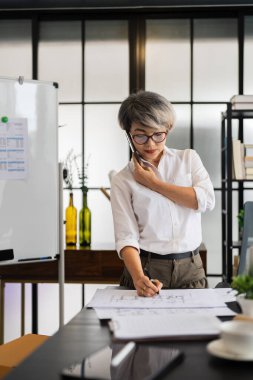 Bir kadın mimar, masasında proje planlarını incelerken, modern ve organize bir çalışma alanında tasarım materyalleri ile çevrili bir telefon görüşmesi yapıyor..