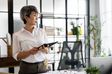 Bir kadın mimar, tasarım kitapları ve materyallerle dolu modern bir ofiste fikirlerini sunmak için özenle bir tablet tutuyor..