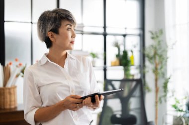 Bir kadın mimar, tasarım kitapları ve materyallerle dolu modern bir ofiste fikirlerini sunmak için özenle bir tablet tutuyor..