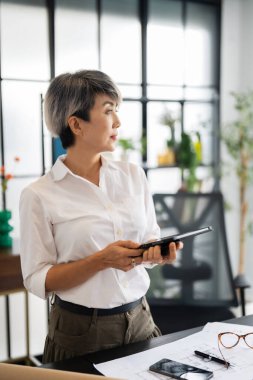 Bir kadın mimar, tasarım kitapları ve materyallerle dolu modern bir ofiste fikirlerini sunmak için özenle bir tablet tutuyor..